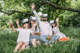 Young caucasian family having fun in virtual glasses on fresh air. Happy parents with two daughters sitting on soft blanket and playing games. Leisure time and entertainment concept.