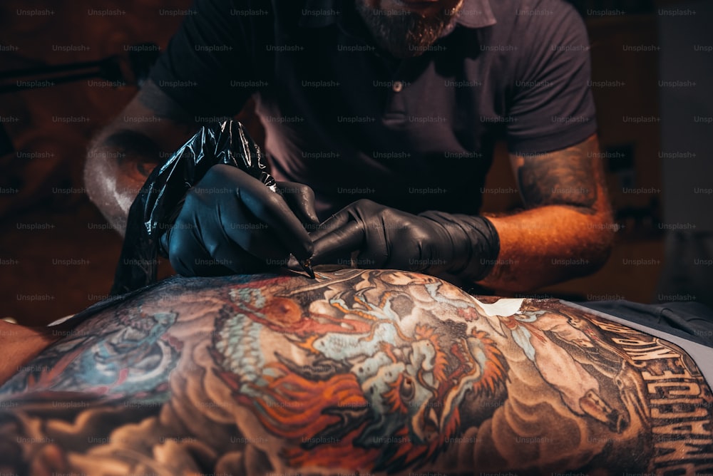 Mãos de tatuador usando luvas de proteção pretas e segurando uma máquina enquanto cria uma imagem nas costas de um homem