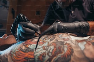 Mãos de tatuador usando luvas de proteção pretas e segurando uma máquina enquanto criam uma imagem nas costas de um homem e a tinta está pingando