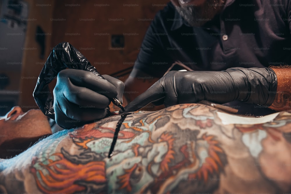 Tätowiererhände, die schwarze Schutzhandschuhe tragen und eine Maschine halten, während sie ein Bild auf dem Rücken eines Mannes erstellen und die Tinte tropft