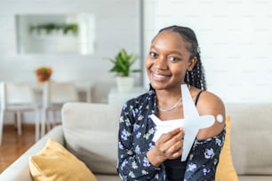 African muslim female smiling after getting a vaccine, holding a paper airplane as a symbol for ready to travel. Woman showing her arm with bandage after receiving vaccination.