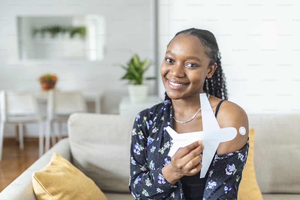 Une femme musulmane africaine souriant après avoir reçu un vaccin, tenant un avion en papier comme symbole de prêt à voyager. Femme montrant son bras avec un bandage après avoir reçu la vaccination.