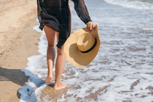 Summer vacation. Carefree woman holding hat and walking on sunny beach at sea waves, close up. Stylish young fit female with straw hat in hands relaxing on tropical shore. Space for text