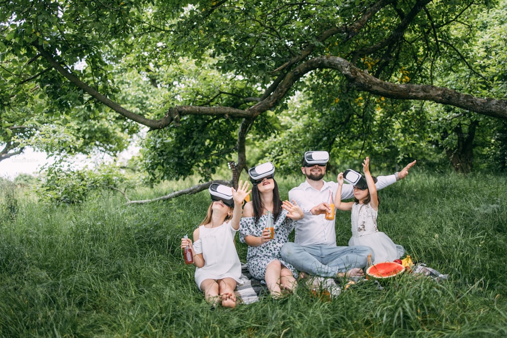 Young caucasian family having fun in virtual glasses on fresh air. Happy parents with two daughters sitting on soft blanket and playing games. Leisure time and entertainment concept.
