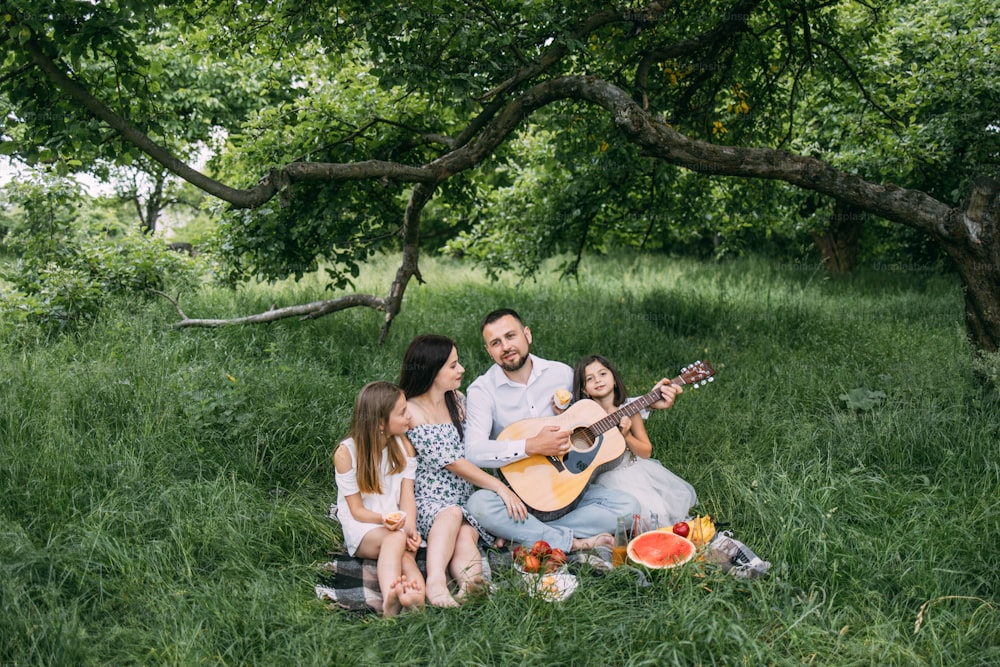 Handsome man playing guitar for his charming wife and two cute daughters. Happy caucasian family sitting on soft blanket at garden and singing songs.
