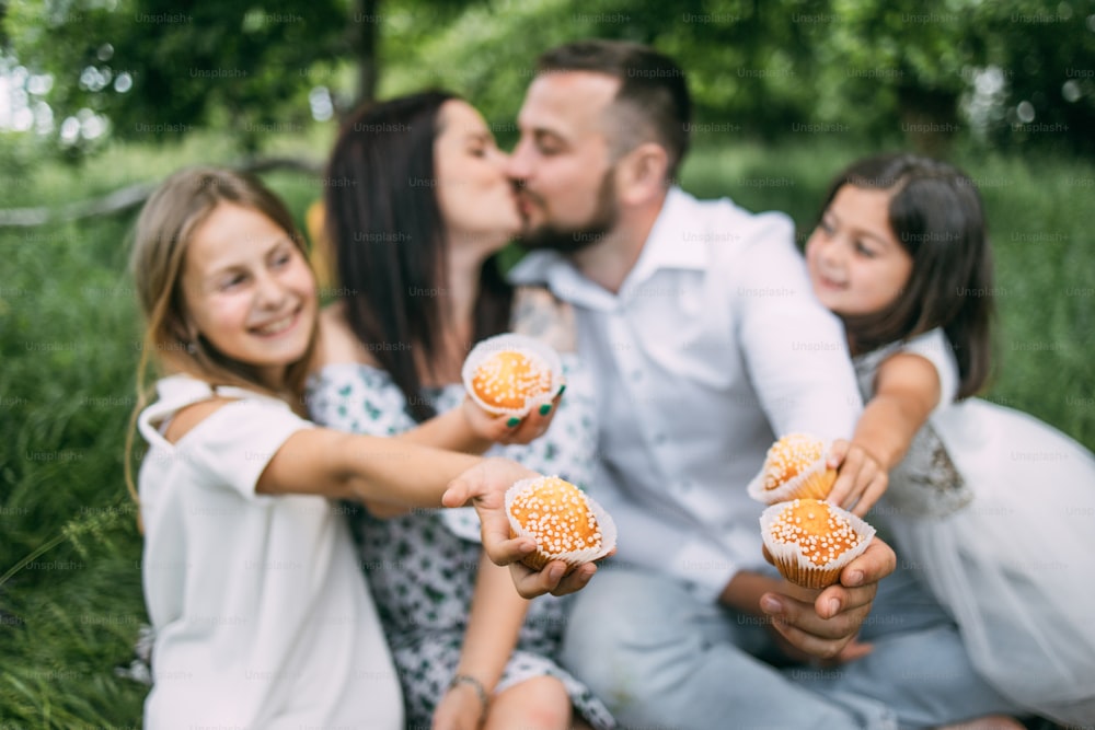 Joyful cute sisters eating delicious cupcakes while father and mother kissing on background. Young family spending leisure time together outdoors. Summer picnic at garden.