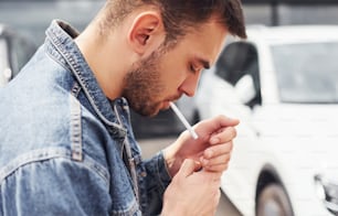 Fashionable man smoking near car outdoors against modern business building.