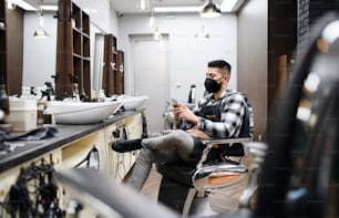 A young man haidresser using smartphone in barber shop, coronavirus and new normal concept.
