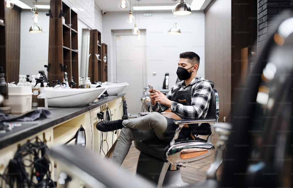 Un giovane parrucchiere che usa lo smartphone nel negozio di barbiere, coronavirus e concetto di nuova normalità.