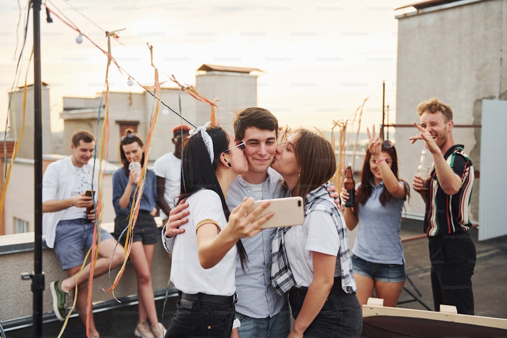 Dos chicas besando a un hombre. Haciéndose un selfie. Un grupo de jóvenes con ropa informal tienen una fiesta juntos en la azotea durante el día.