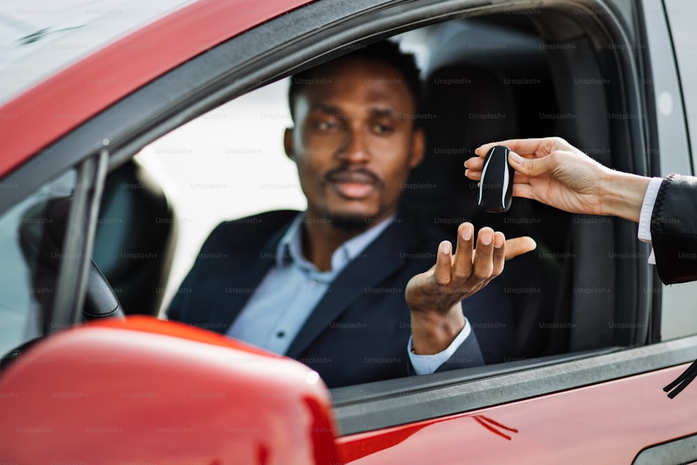新しい電気自動車の男性客に鍵を渡す女性自動車販売業者。高級な赤い自動車の中に座っているスーツのハンサムなアフリカの男性。手に焦点を合わせます。