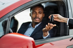 Female auto seller giving keys to male customer form his new electric car. Handsome african man in suit sitting inside luxury red auto. Focus on hands.