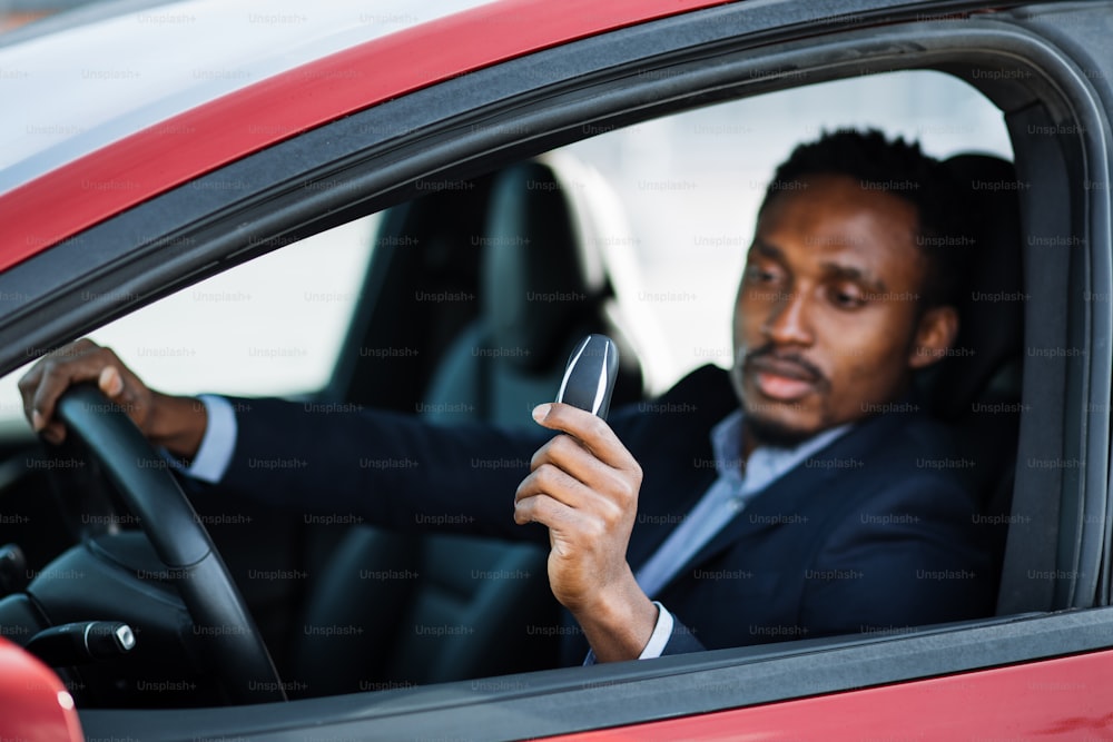 Empresario afroamericano de traje sentado dentro de su lujoso coche eléctrico con las llaves en la mano. El hombre guapo parece satisfecho con una compra exitosa. Concepto de vehículo ecológico.