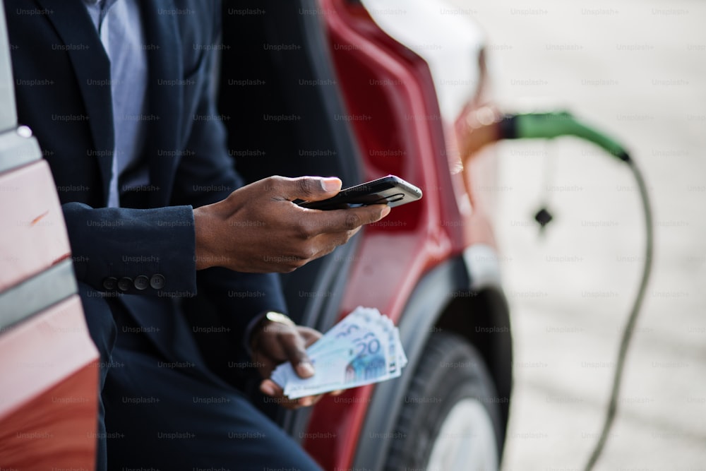 Uomo d'affari africano serio seduto in auto rossa con portiera aperta e in possesso di smartphone con denaro contante. L'auto di lusso è in carica. Concetto di risparmio.