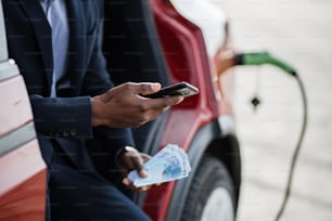 Seriöser afrikanischer Geschäftsmann sitzt in einem roten Auto mit geöffneter Tür und hält ein Smartphone mit Bargeld in der Hand. Luxusauto wird aufgeladen. Konzept der Einsparungen.