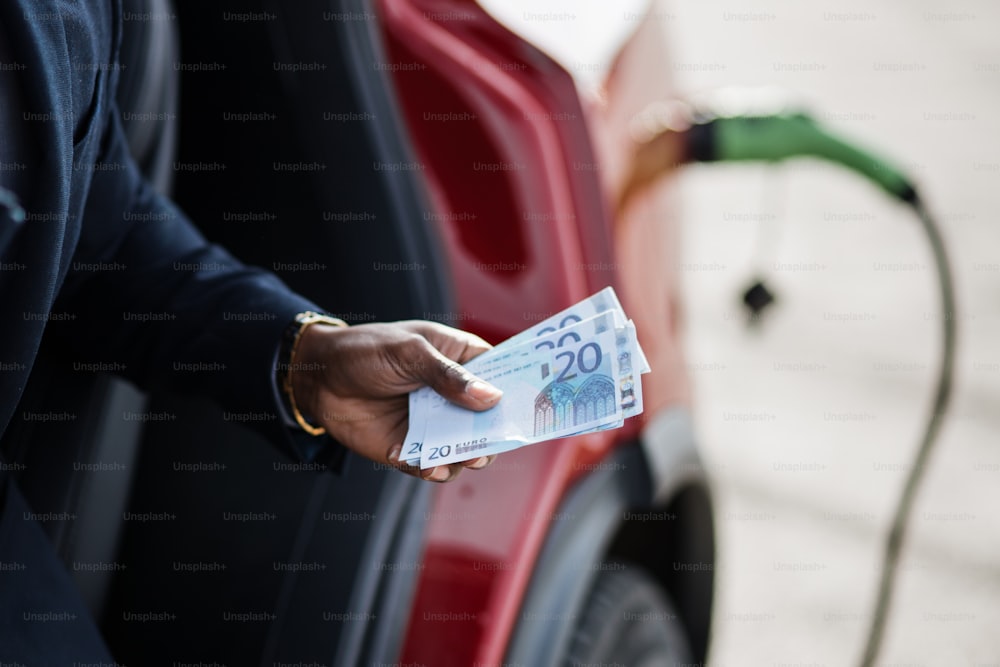 Close up da mão masculina segurando dinheiro dinheiro sobre fundo desfocado de carregamento de carro elétrico. Empresário africano inteligente de terno economizando dinheiro em veículo ecológico.