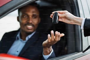 Female auto seller giving keys to male customer form his new electric car. Handsome african man in suit sitting inside luxury red auto. Focus on hands.