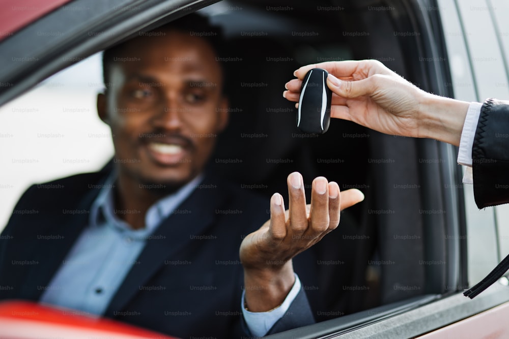 Female auto seller giving keys to male customer form his new electric car. Handsome african man in suit sitting inside luxury red auto. Focus on hands.