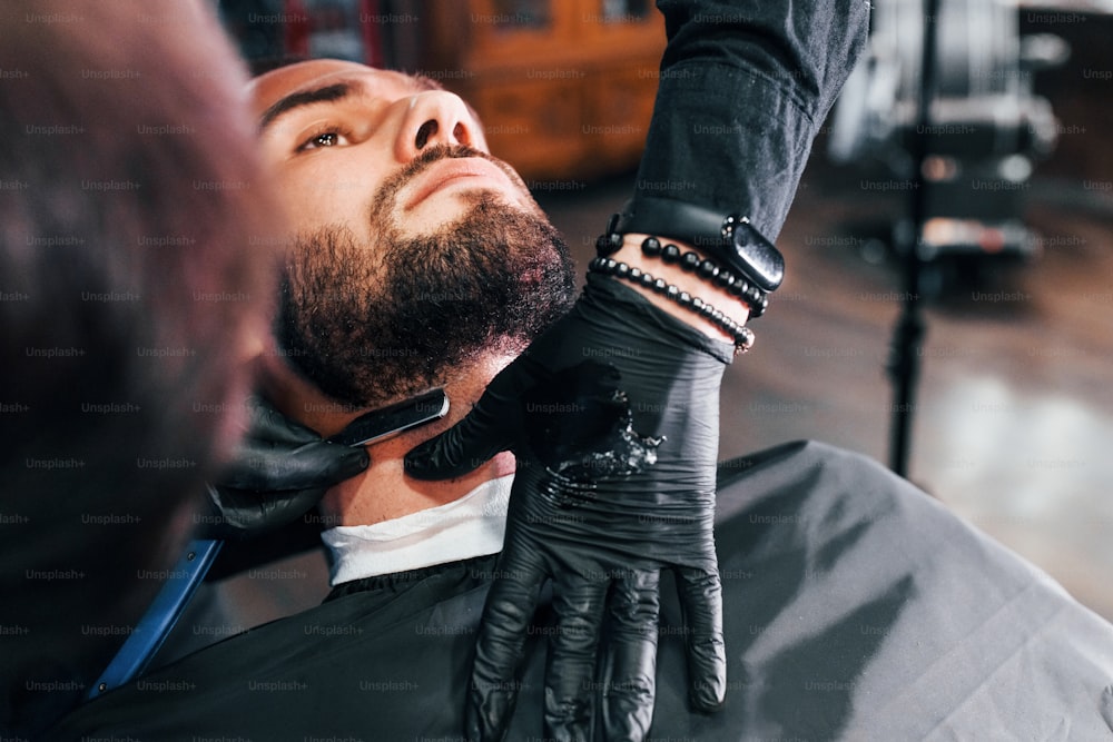 Young man with stylish hairstyle sitting and getting his beard shaved in barber shop.