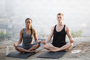 Young multicultural couple in sport outfit sitting in lotus position and meditation on yoga mat outdoors. Concept of breathing practice and peacefulness.