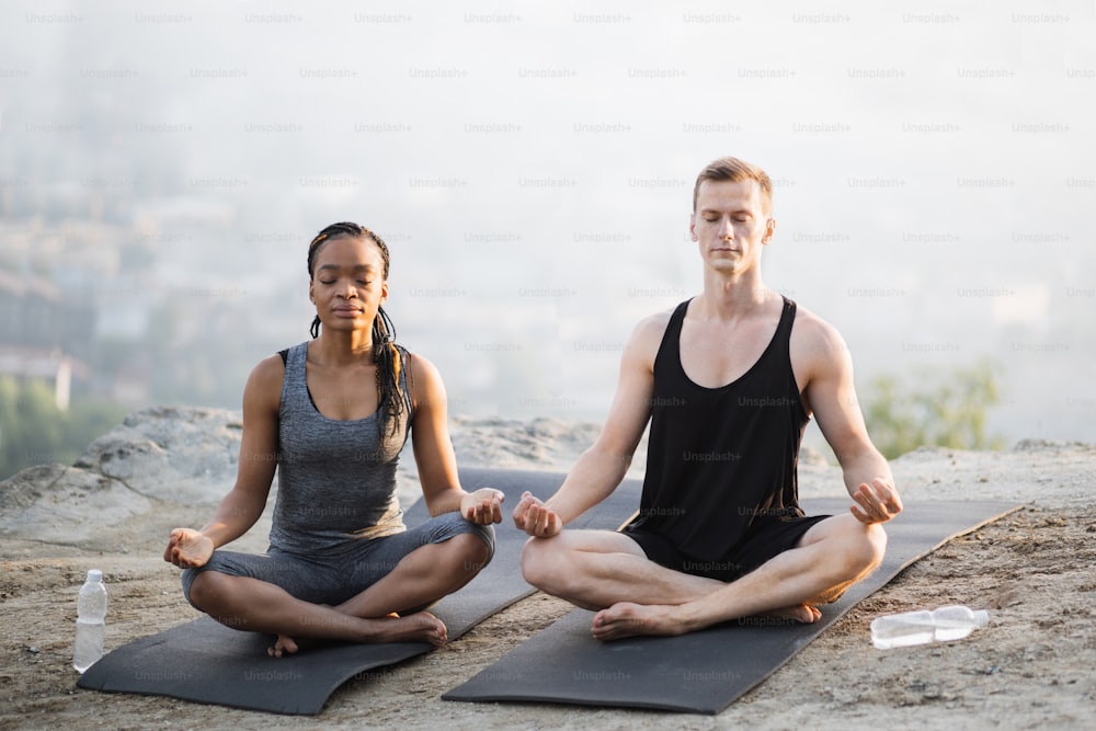 Giovane coppia multiculturale in tenuta sportiva seduta in posizione di loto e meditazione sul tappetino da yoga all'aperto. Concetto di pratica respiratoria e tranquillità.