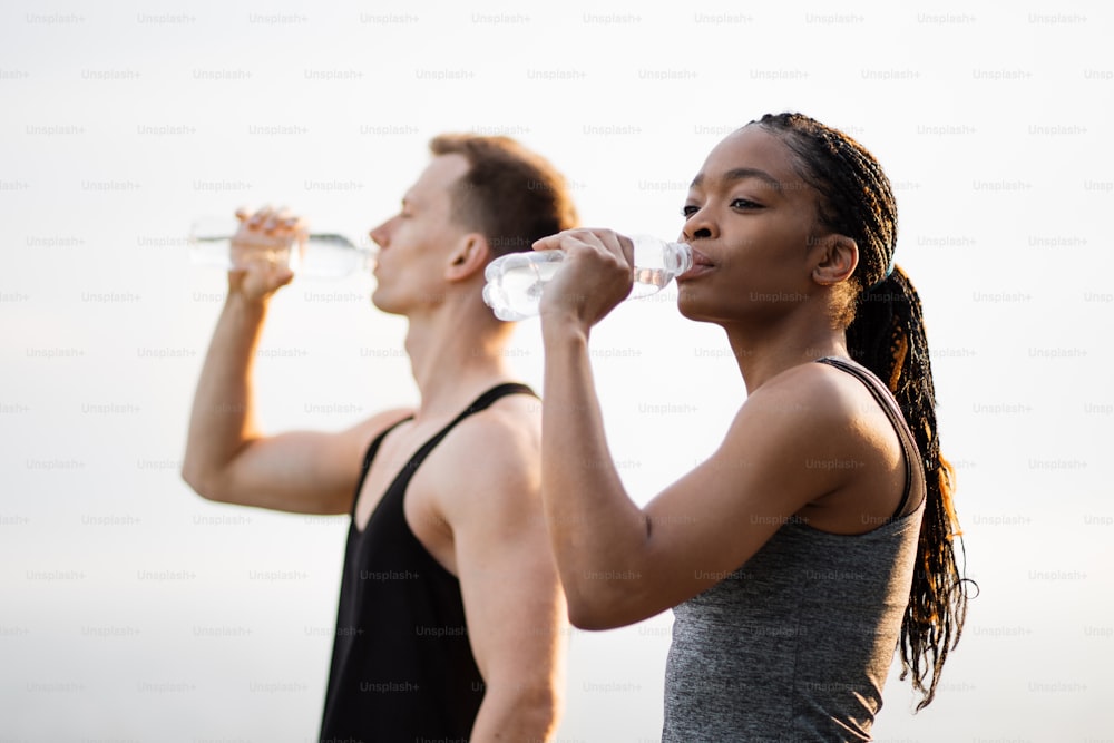 Side view of african american woman and caucasian man refreshing with cold water after active outdoors workout. Concept of people,healthy lifestyles and recreation.