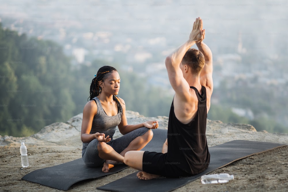 Pareja joven multiétnica sentada cara a cara en esterilla y practicando yoga al aire libre. Hombre y mujer deportistas y saludables que disfrutan de un entrenamiento regular al aire libre.