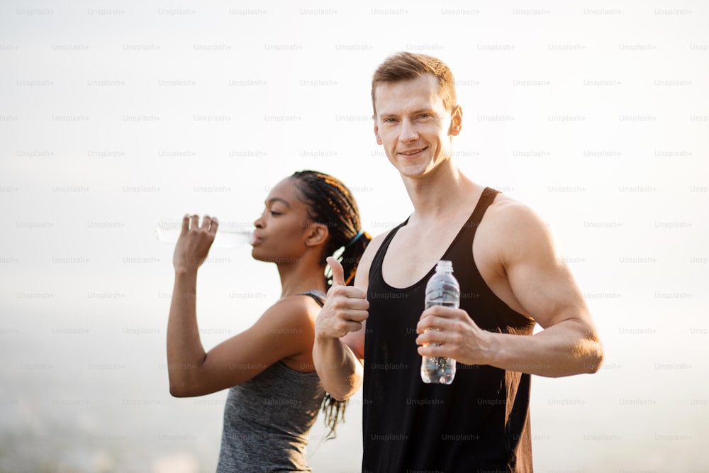 Multiethnic couple in sportswear standing outdoors and taking break during workout. African woman drinking water, caucasian man showing thumb up.