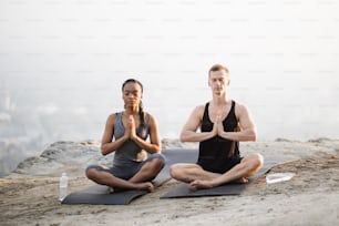 Peaceful multi ethnic couple in sport clothes sitting in lotus position yoga mat and with namaste hands. Young man and woman keeping eyes closed while meditating on fresh air.