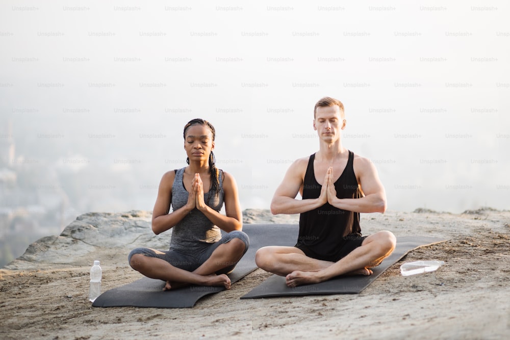 Friedliches multiethnisches Paar in Sportkleidung sitzt in Lotusposition Yogamatte und mit Namaste-Händen. Junger Mann und junge Frau, die die Augen geschlossen halten, während sie an der frischen Luft meditieren.