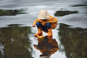 Kind in gelbem wasserdichtem Umhang und Stiefeln spielt mit handgemachtem Bootsspielzeug aus Papier im Freien nach dem Regen.