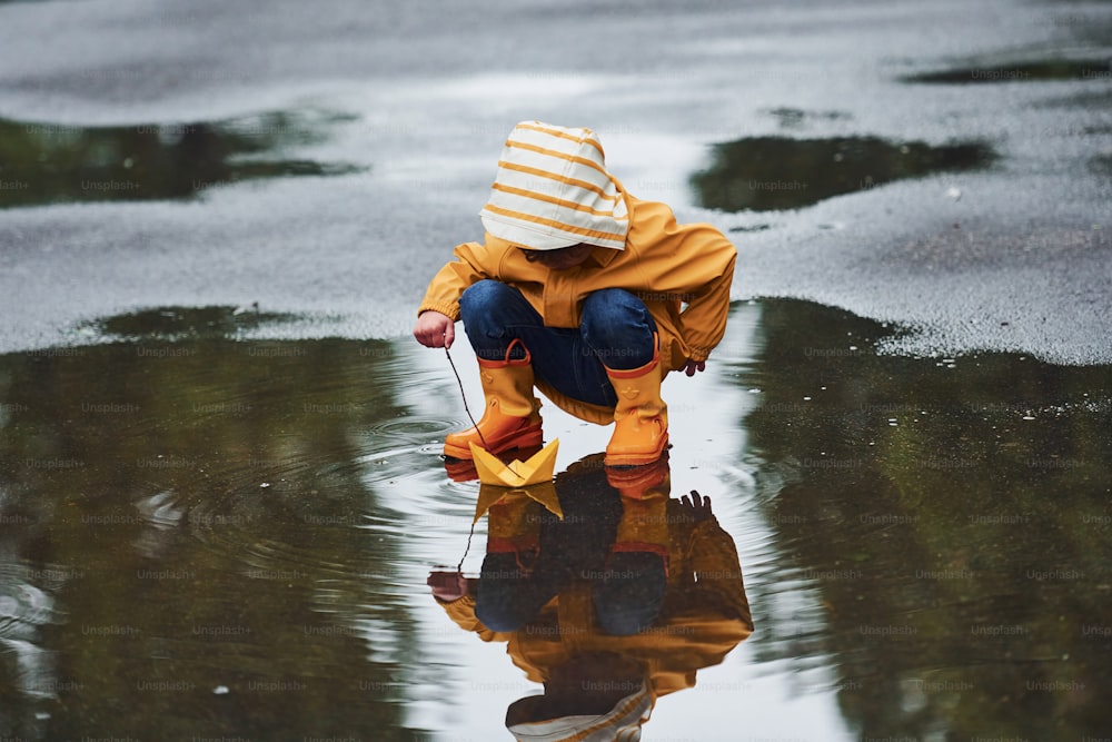Kid in yellow waterproof cloak and boots playing with paper handmade boat toy outdoors after the rain.