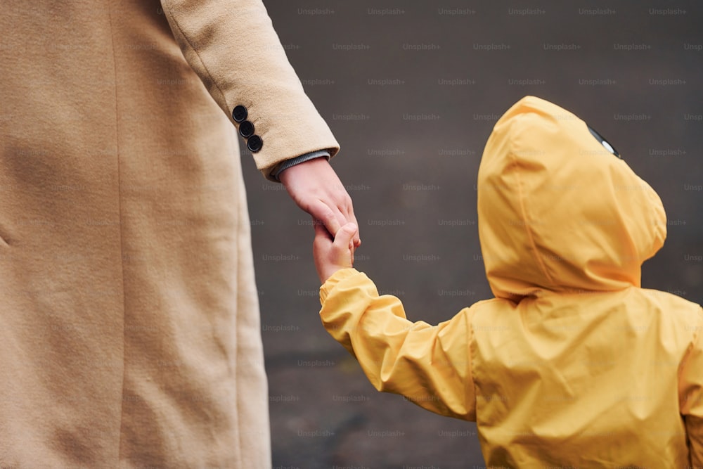 黄色い防水マントを着て母親の手を持ち、屋外を散歩する子供。