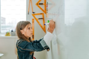 Estudante do ensino fundamental escrevendo no quadro-negro por feltro-tip na sala de aula da escola moderna. Conceito de volta às aulas. Foco seletivo.