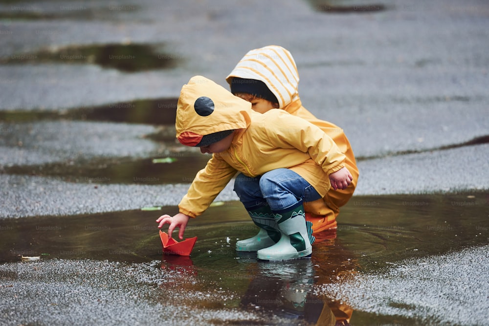 Dos niños con capas amarillas impermeables y botas jugando juntos con un bote de papel hecho a mano al aire libre después de la lluvia.