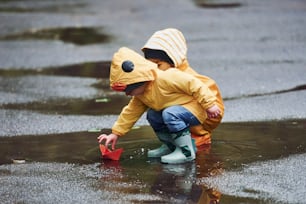 Dos niños con capas amarillas impermeables y botas jugando juntos con un bote de papel hecho a mano al aire libre después de la lluvia.