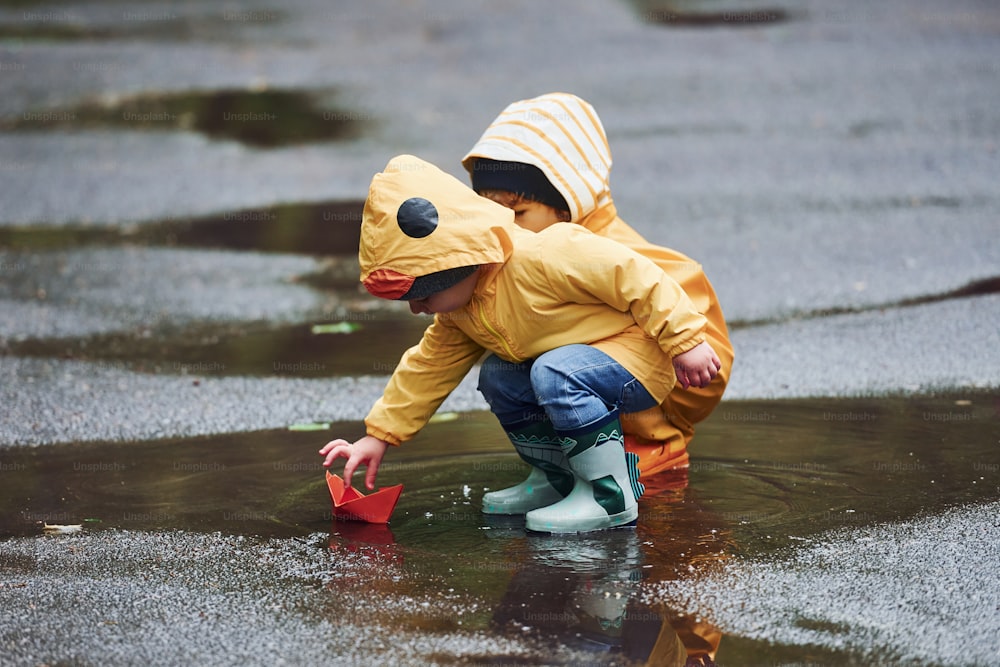 黄色い防水マントとブーツを身に着けた2人の子供が、雨上がりに屋外で手作りの紙のボートで遊んでいます。