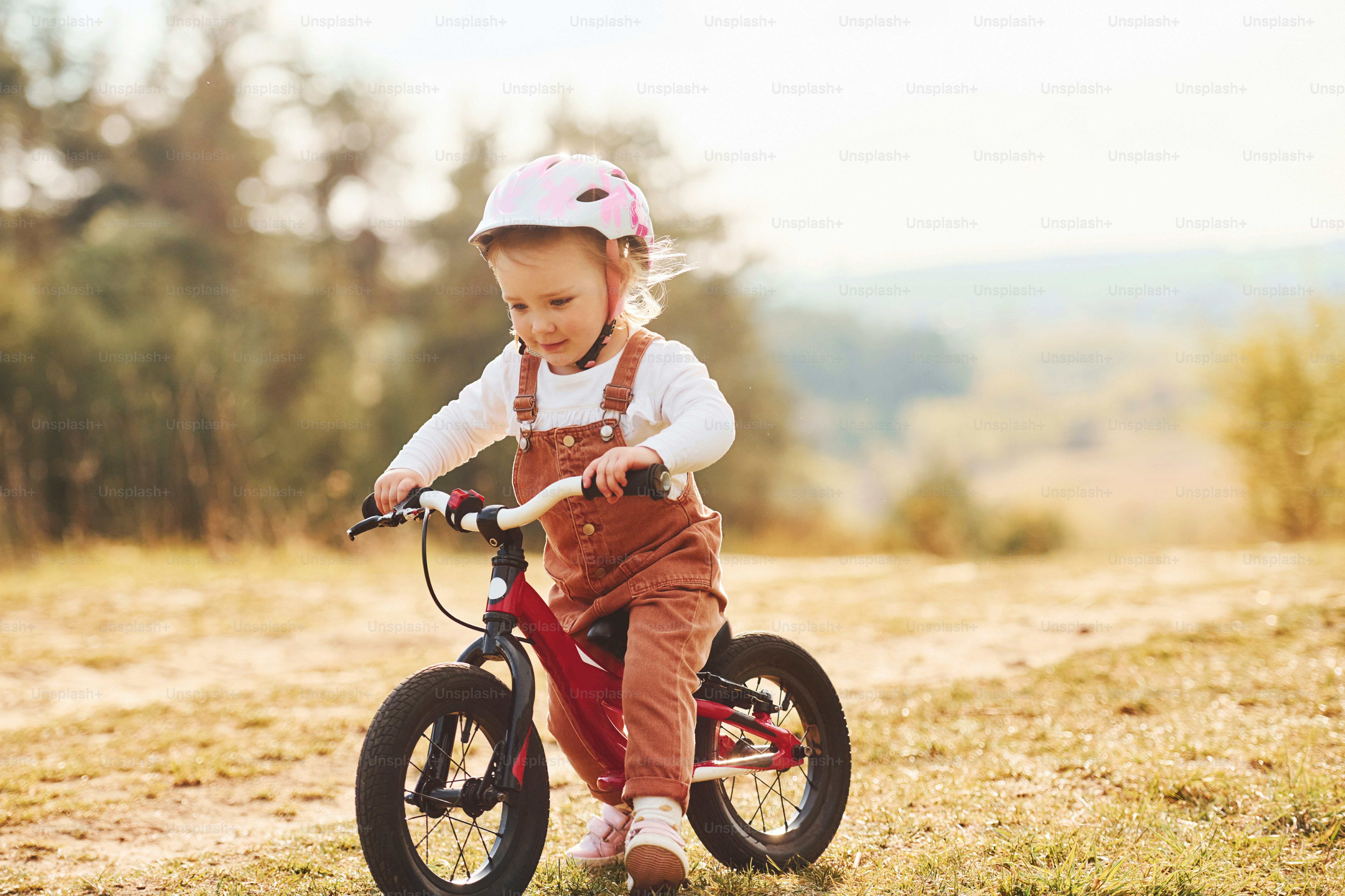 Happy little girl in protective hat riding her bike outdoors at sunny day near forest.