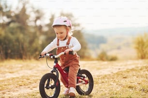 Happy little girl in protective hat riding her bike outdoors at sunny day near forest.