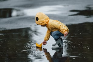 Bambino in mantello impermeabile giallo e stivali che gioca con il giocattolo della barca fatto a mano di carta all'aperto dopo la pioggia.