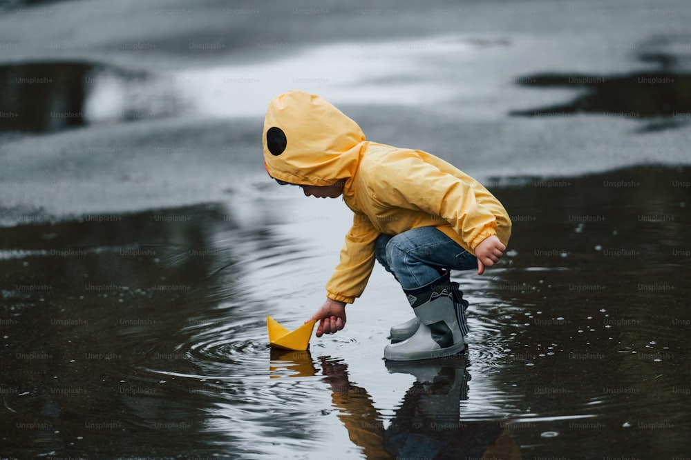 Kid in yellow waterproof cloak and boots playing with paper handmade boat toy outdoors after the rain.