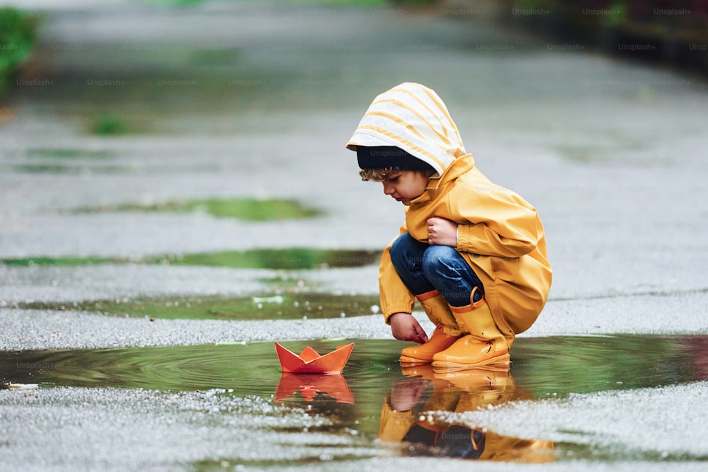 Kind in gelbem wasserdichtem Umhang und Stiefeln spielt mit handgemachtem Bootsspielzeug aus Papier im Freien nach dem Regen.