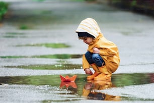 Kid in yellow waterproof cloak and boots playing with paper handmade boat toy outdoors after the rain.