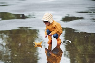 Kid in yellow waterproof cloak and boots playing with paper handmade boat toy outdoors after the rain.