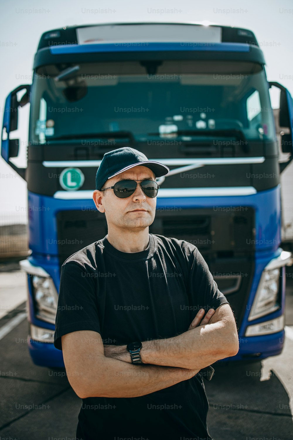 Professional truck driver with hat and sunglasses confidently standing in front of big and modern truck. Bright sunny day. People and transportation concept.
