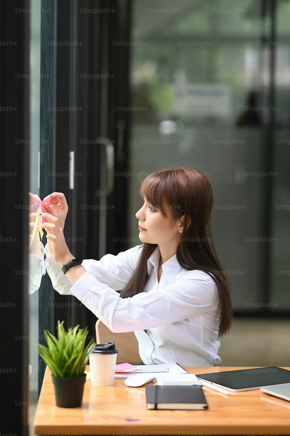 Female office worker putting sticky note on glass wall at modern office.