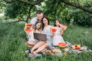 Joyful caucasian family having summer picnic with sweet fruits and cupcakes. Woman holding wireless laptop while two daughters and husband waving while having video call. Happiness concept.