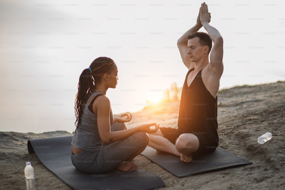 Relaxed multicultural couple meditating with closed eyes on yoga mat during summer sunset on high rock. Concept of people, healthy and active lifestyles.