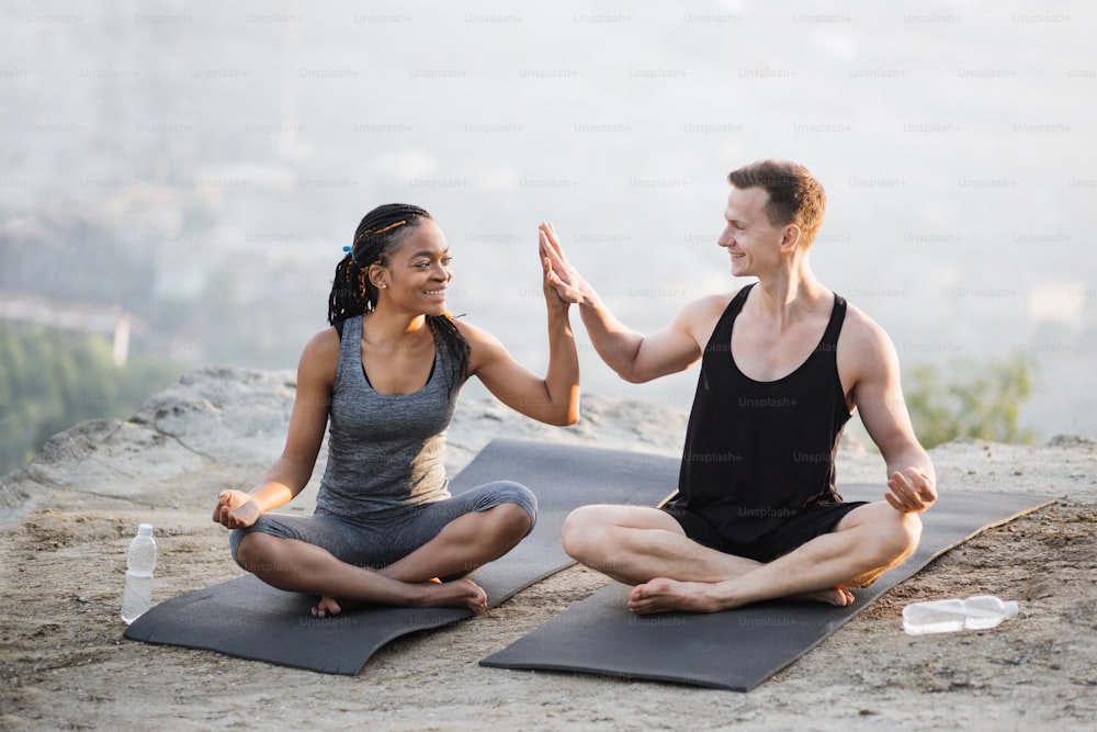 Sporty healthy couple sitting on yoga mat in lotus position and giving high five during workout on fresh air. Caucasian man and african woman enjoying outdoors training together.