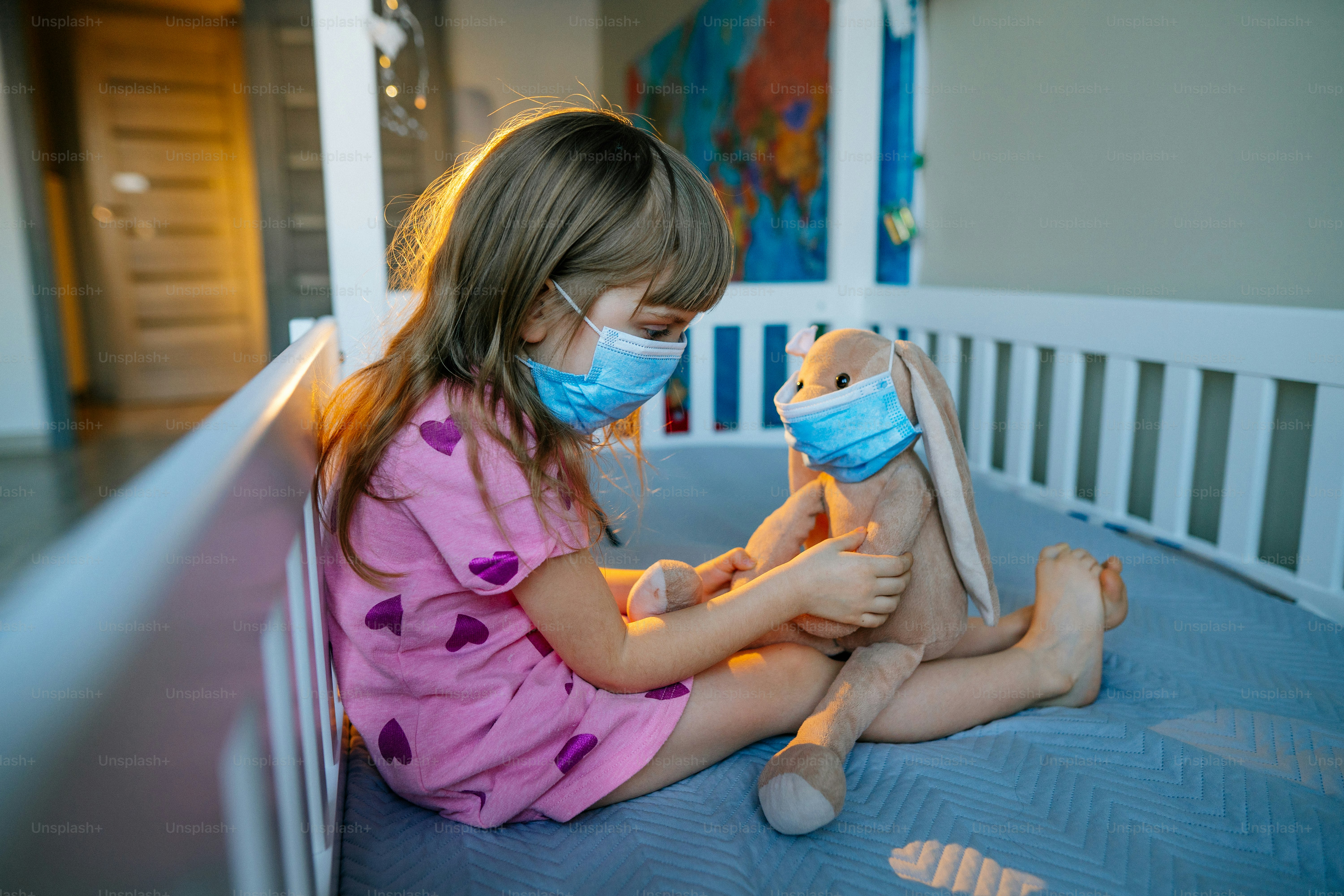 Kleines 4-jähriges Mädchen mit Schutzmaske spielt mit ihrem Plüschtierhasen, das auf dem Bett im Kinderzimmer sitzt. Neue Normalität nach pandemischem COVID-19-Konzept.